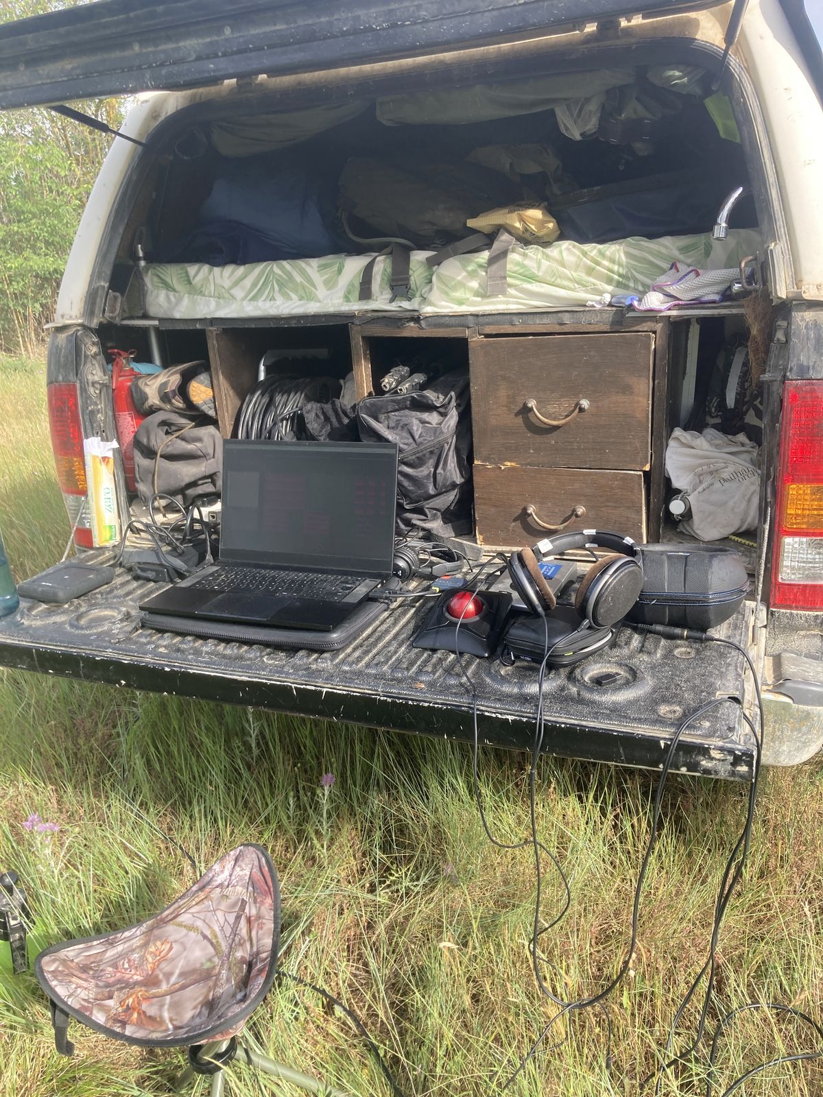 A laptop and headphones on the open tailgate of the truck, in long grass
