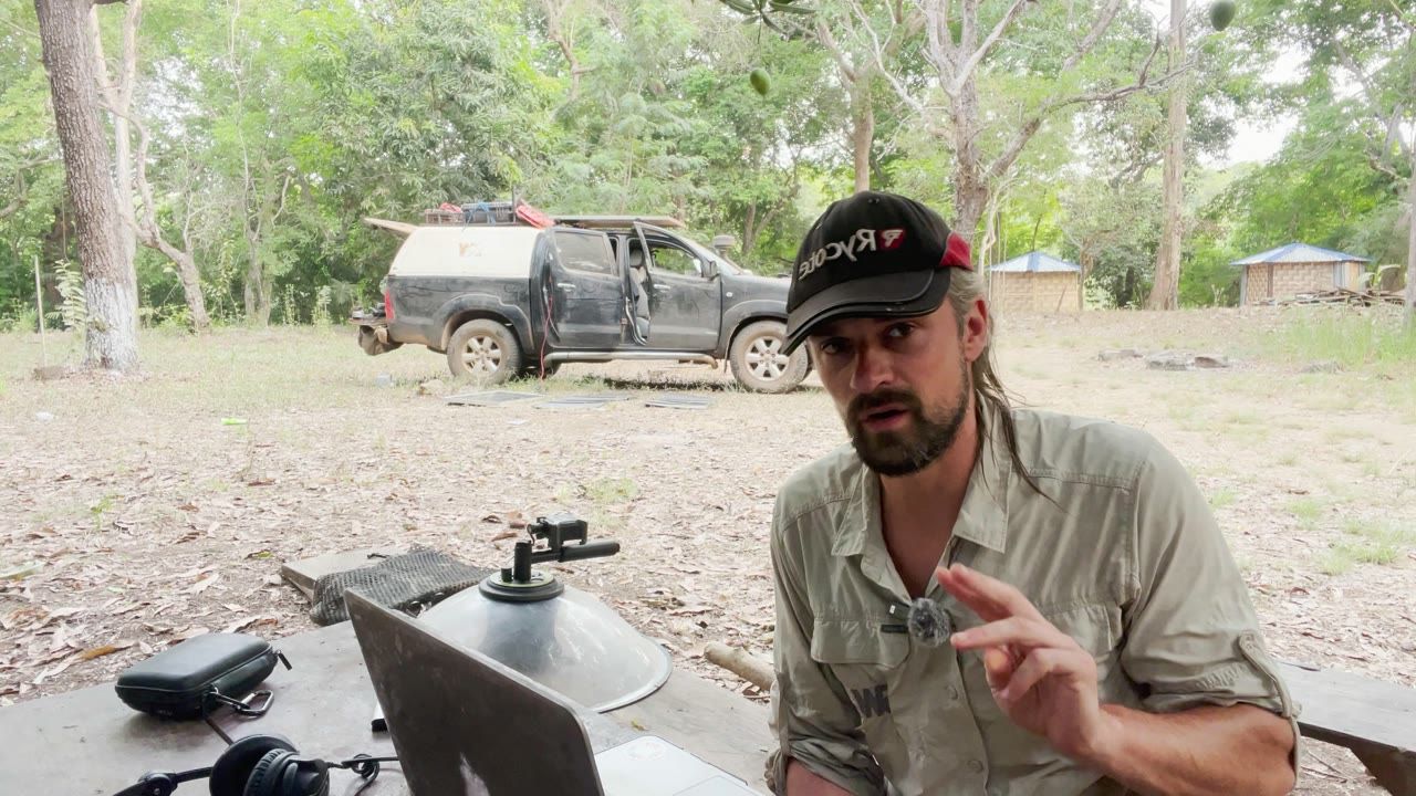 Axel at a camp table with a laptop and recorder, his pickup truck behind him among the trees