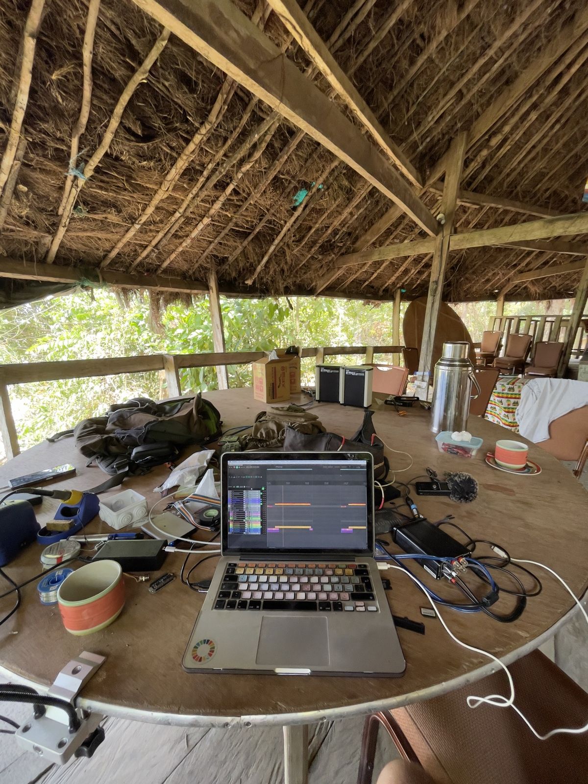 A laptop showing an audio session on a round table under a thatched roof, with recorders and cables